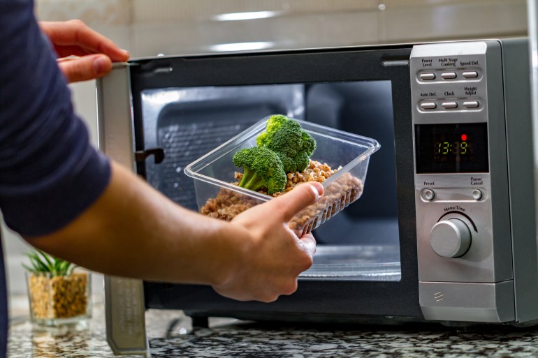 Using the microwave oven to heat food. Woman's hand puts plastic container with broccoli and buckwheat in the microwave