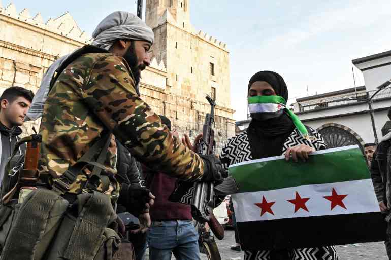 A man hands over an assault rifle to a masked woman holding a sign depicting the Syrian opposition flag and wearing the same flag across her face, outside the Umayyad Mosque in Damascus on December 10, 2024. Islamist-led rebels took Damascus in a lightning offensive on December 8, ousting president Bashar al-Assad and ending five decades of Baath rule in Syria. (Photo by LOUAI BESHARA / AFP)